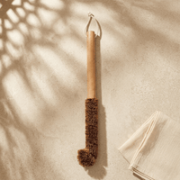 Natural coconut fibre bottle cleaning brush with wooden handle, hanging in a minimal room setting.