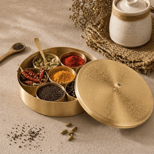 Open brass masala dabba with seven spice containers and lid beside